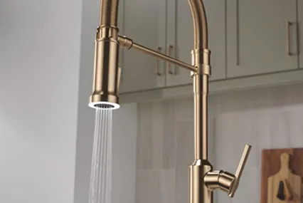 Single handle Delta Spotlight Task Light Faucet shown in champagne bronze. Water pours out. Kitchen wall with cabinets in background.