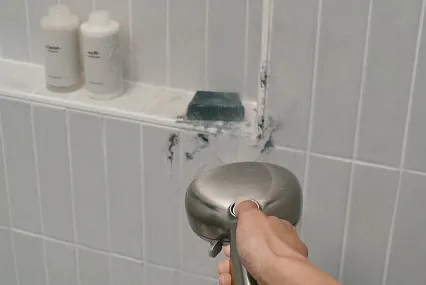 Delta SpotShield Brushed Nickel shower head in hand seen from viewer’s perspective using ProClean to spray off soap scum on shower shelf.
