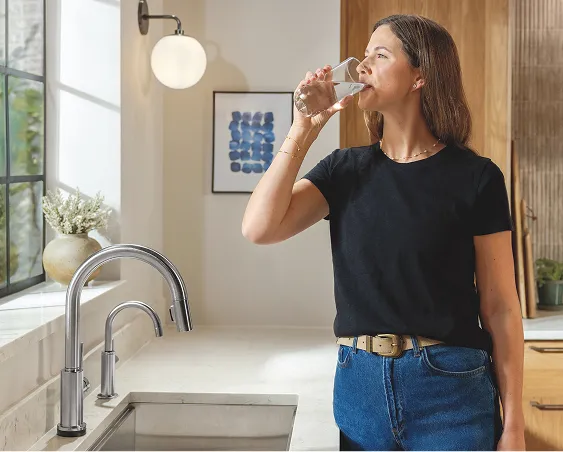 Woman standing in front of sink with chrome Delta kitchen faucets enjoys glass of water while looking out the window.