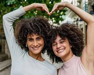 Two women with curly hair stand together and form a heart shape with their arms