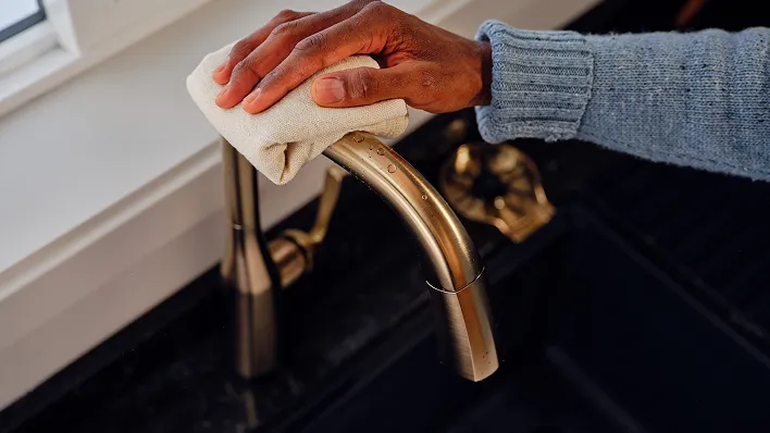 Hand with cloth wipes the top of a Delta single handle kitchen faucet in champagne bronze with Lumicoat finish.