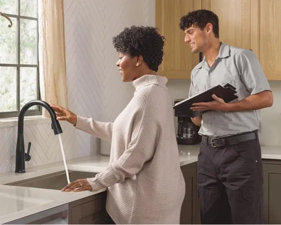 Smiling woman stands at kitchen sink admiring new matte black Delta single-handle faucet while Delta installer looks on proudly