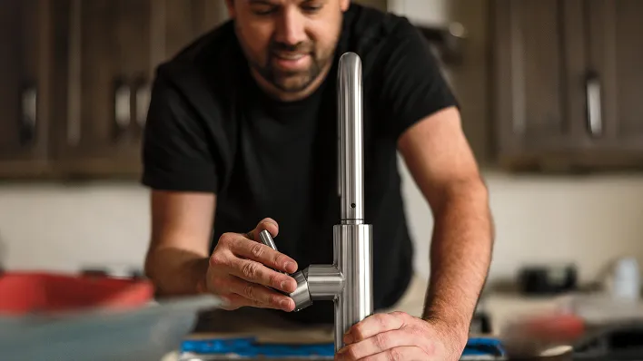 Plumber/installer in kitchen with dark cabinets (background) installing chrome single-handle Delta faucet (foreground).
