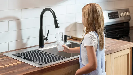 Girl stands in front of sink and fills glass of water from Delta Essa Touch2O Technology Kitchen Faucet with Touchless Technology in Matte Black.
