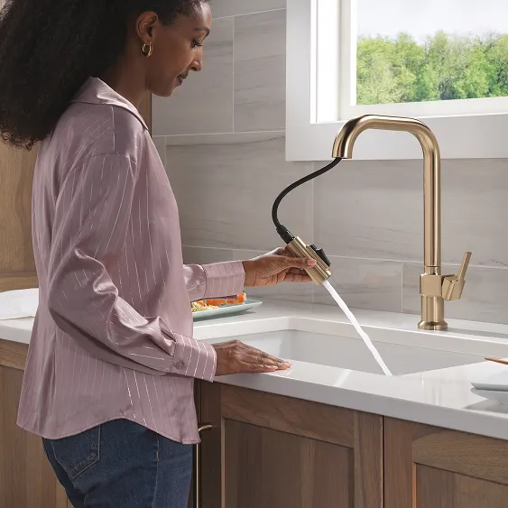 Woman stands at white kitchen counter with grey backsplash and uses pull-out nozzle from Delta Noreau Kitchen Faucet in Champagne Bronze to spray into undermount sink.
